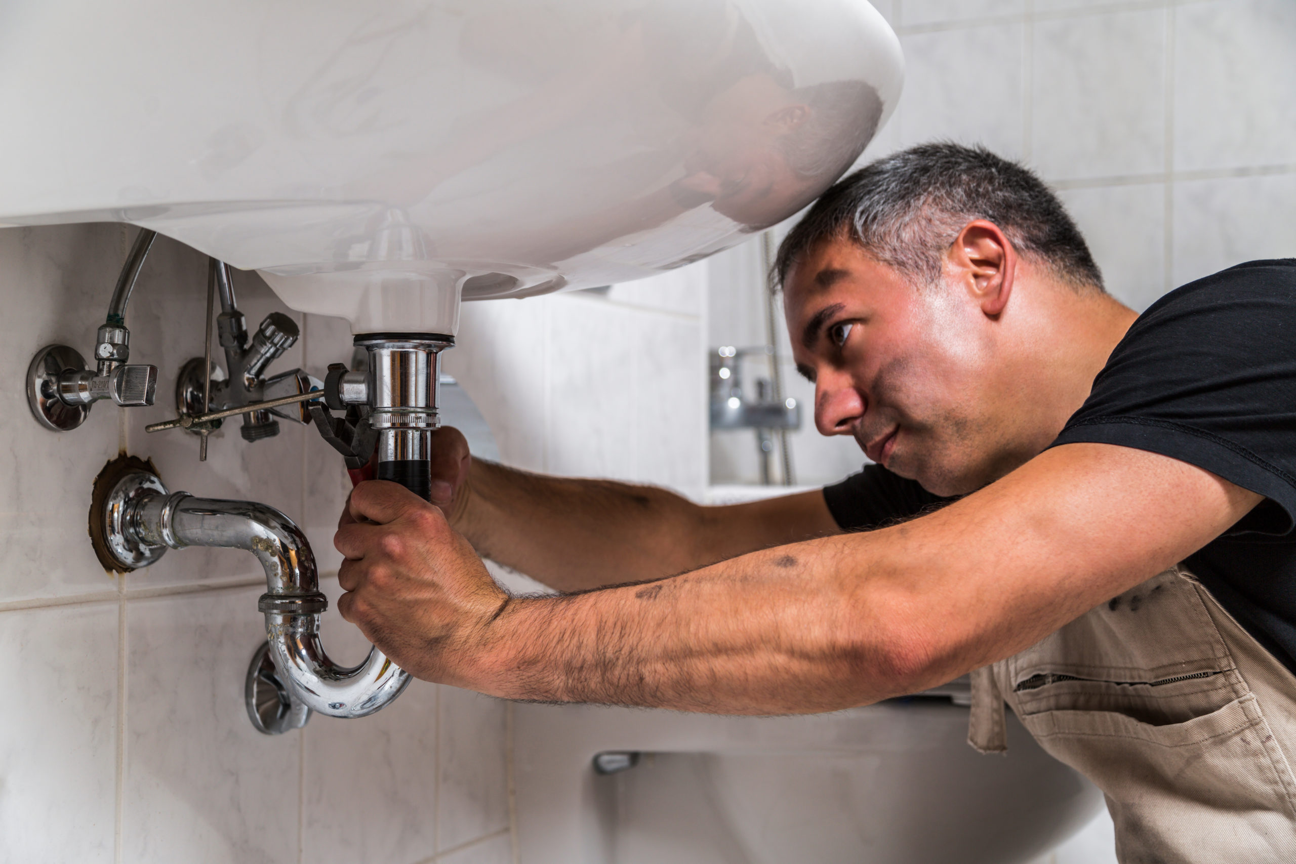 male man dirty muscle inflated worker specialist plumber in black t-shirt and white old shabby working suit repairs faucet pipe with a metallic pliers in bathroom.